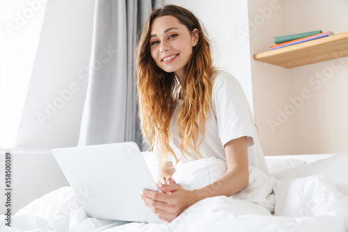 Portrait of ginger joyful woman using laptop while sitting in bed