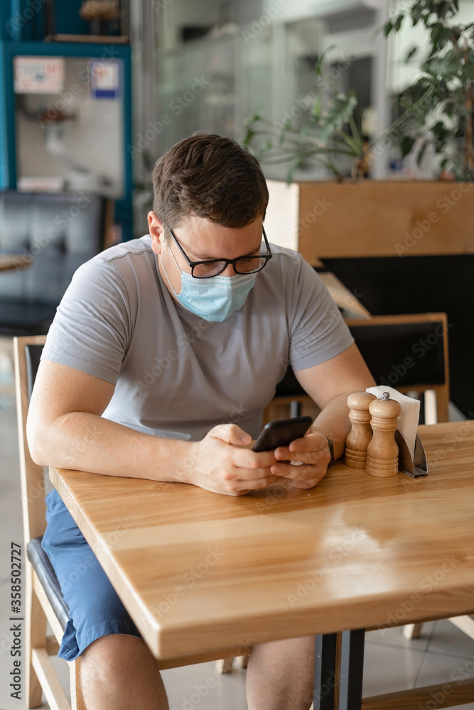 Caucasian man sitting in restaurant and using phone in medical face mask. New Normal concept Caucasian man sitting in restaurant and using phone in medical face mask. New Normal concept