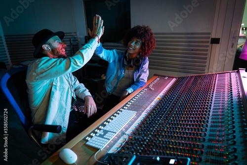 A professional male producer and young african female singer are happy with results of recording a new song and  are celebrating a future success in a music studio with colorful lights on a background