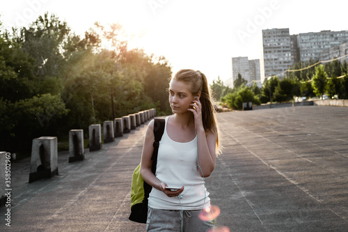 Beauty young female model with long blond hairstyle in sport clothes and with backpack standing on the road and looking sideways. The girl listen to music in earphones and walking on the cort photo