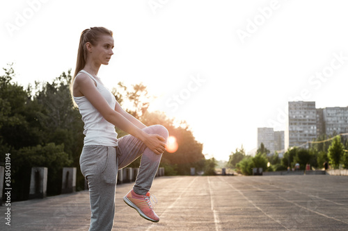 Girl stretching her legs outdoor and preparing to sport trainings. Female model posing outside on air in sporty clothes and doing exercises © darakaliton