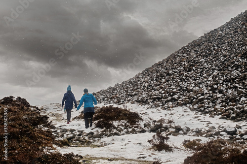 Two women walking in a mountain in a snow fall by burial chamber of Queen Maebh of Connacht county Knocknarea hill, Sligo, Ireland. Winter scene, photo