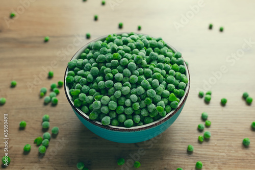 Frozen green peas in a bowl with few scattered on the wooden table
