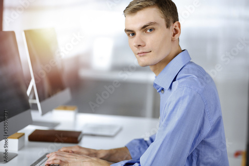 Young successful businessman works with computer  sitting at the desk in a sunny modern office. Headshot of male entrepreneur or it-specialist at workplace. Business concept