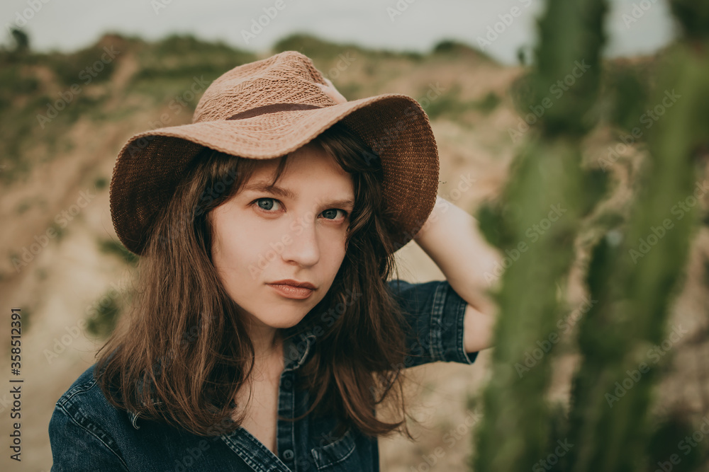 Young beautiful woman sitting on the sand in a blue denim suit and red hat in a Western style in a quarry with cacti. The idea of shooting a wild west.