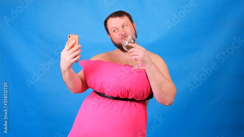 Funny bearded guy in a pink dress made of pillows with a glass of wine makes selfie on a blue background. Crazy quarantine. Funny house cleaning. Fashion 2020. Put on a pillow. Challenge 2020 due to © Евгения Медведева