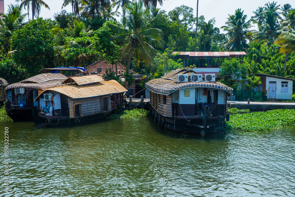 Small houses in a local village located next to Kerala's backwater on a bright sunny day and traditional Houseboat seen sailing through the picturesque backwaters of Allapuzza or Alleppey in Kerala Small houses in a local village located next to Kerala's backwater on a bright sunny day and traditional Houseboat seen sailing through the picturesque backwaters of Allapuzza or Alleppey in Kerala