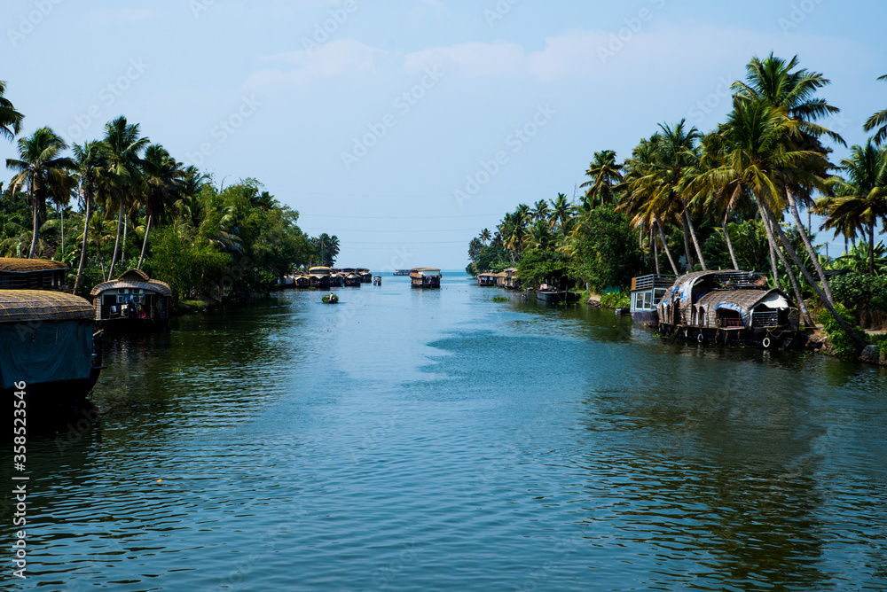 Small houses in a local village located next to Kerala's backwater on a bright sunny day and traditional Houseboat seen sailing through the picturesque backwaters of Allapuzza or Alleppey in Kerala