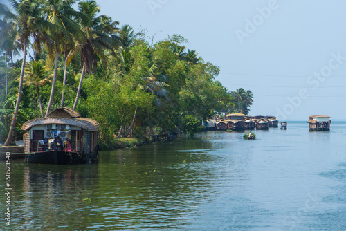 Small houses in a local village located next to Kerala s backwater on a bright sunny day and traditional Houseboat seen sailing through the picturesque backwaters of Allapuzza or Alleppey in Kerala 