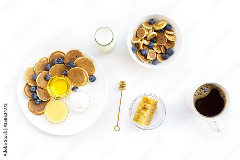Mini Pancake cereal and middle size pancakes with two different honey and honeycomb, yogurt, milk and blueberry on white plate and black coffee on white cup.