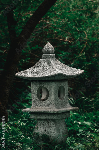 Japanese stone lantern or light tower surrounded by fresh green plants, asian traditional architecture, exotic park ornament in japanese zen garden, Buddhist temple element, relaxing spot in nature 
