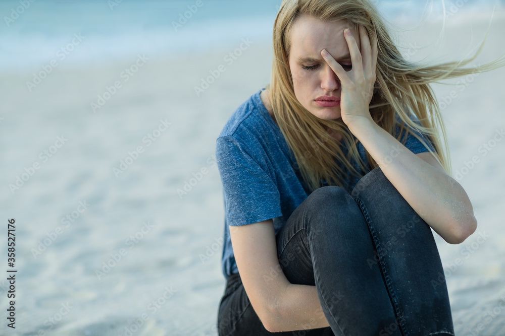 Sad young woman with hand on face sitting alone on the beach. Feeling depressed with mental pain.