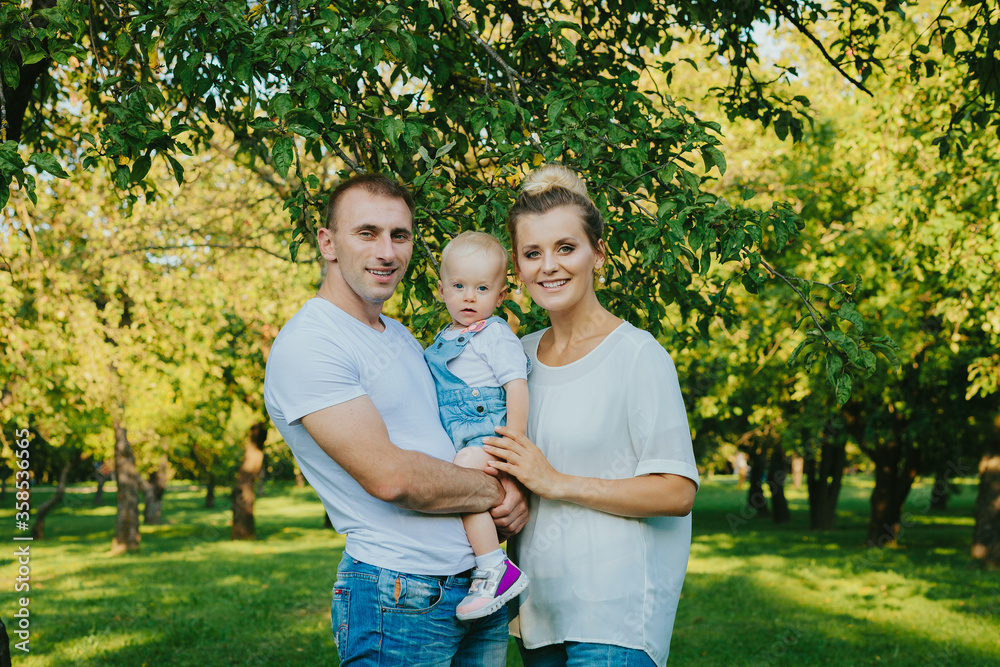 Fototapeta premium Happy young family on nature background. Smiling father and mother holding their little child's in his arms. Young man, woman and child in park on summer. Love and harmony in family.