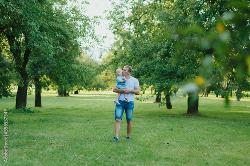 Happy father holds his baby daughter in his arms and kissing her on nature background in summer day. Young man with his little child walking in park on summer. Happy paternity. Father's love. 