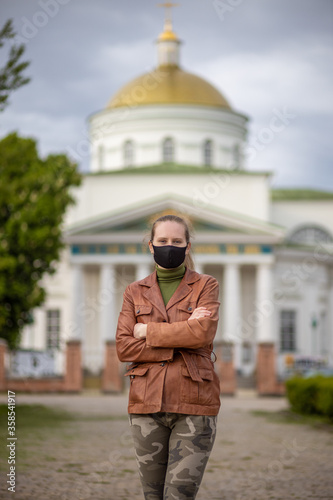 Portrait of a beautiful woman in a black mask, who stands against the backdrop of the Orthodox Church. Woman in a brown jacket and khaki pants.