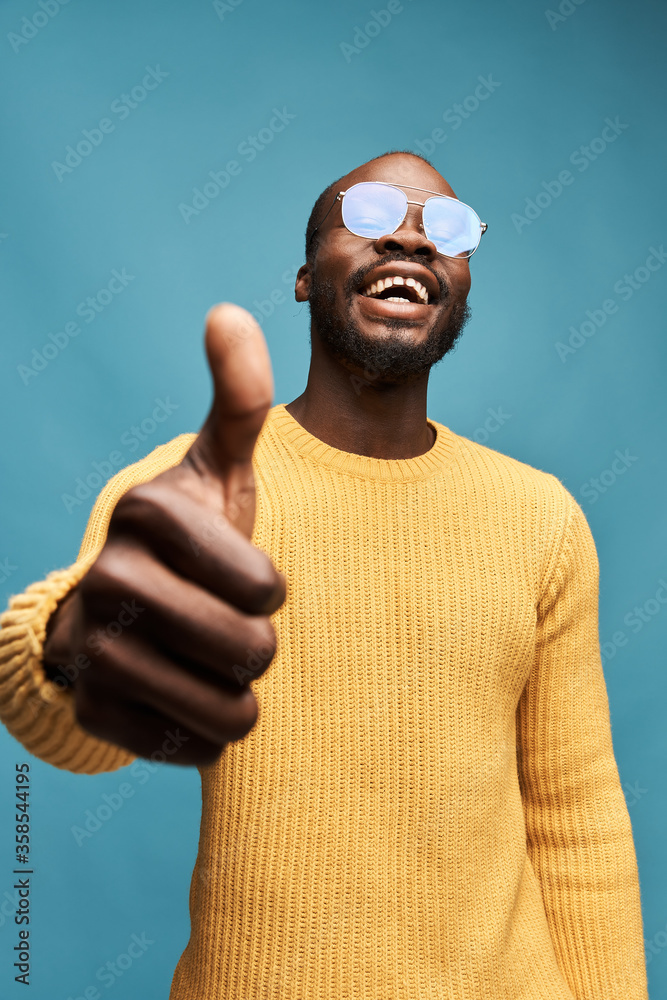 Naklejka premium portrait photo of a dark-skinned handsome guy with a beard on a blue background in a yellow sweater, transparent glasses, he looks at the camera, smiles and keeps his thumb up