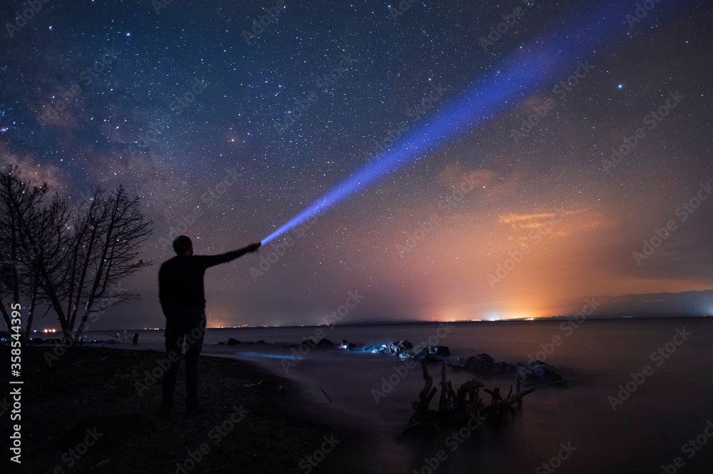 Beautiful starry sky with bright milky way galaxy. Night landscape . Person silhouette stands on the shore with flashlight illuminate starry sky.
