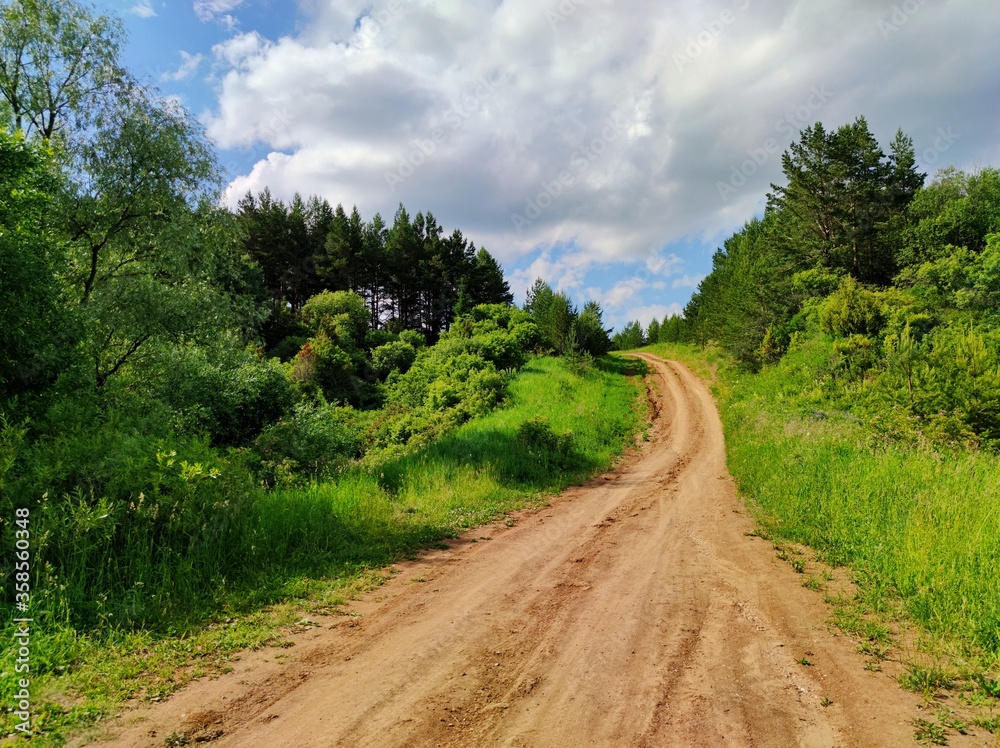 sunny blue sky with clouds over a country road leading uphill among green grass and trees