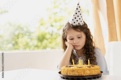 . Girls birthday and birthday cake with candles. The child is sad and laid his head on his raised hand. In the background is a large window. The room is bright and comfortable.