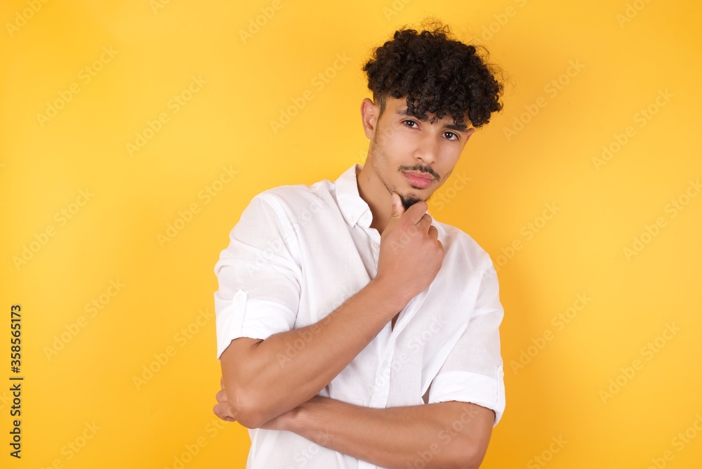 Portrait of thoughtful smiling man keeps hand under chin, looks directly at camera, listens something with interest, dressed casually, poses against gray wall. Youth concept. Portrait of thoughtful smiling man keeps hand under chin, looks directly at camera, listens something with interest, dressed casually, poses against gray wall. Youth concept.