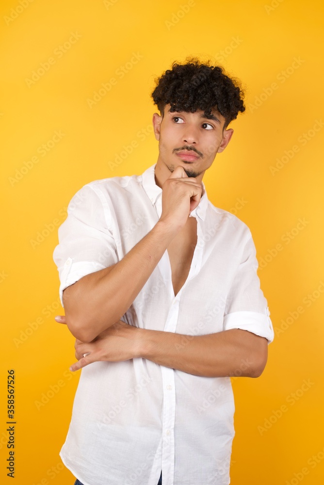 Face expressions and emotions. Thoughtful young man in casual clothes holding hand under his head, having doubtful look.