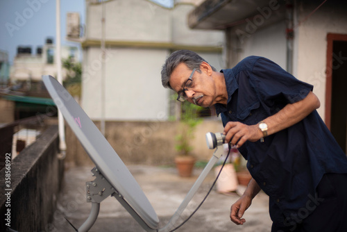 An old/aged Indian Bengali man in blue shirt is trying to fix a dish antenna standing on a rooftop under the open sky. Indian lifestyle and seniors