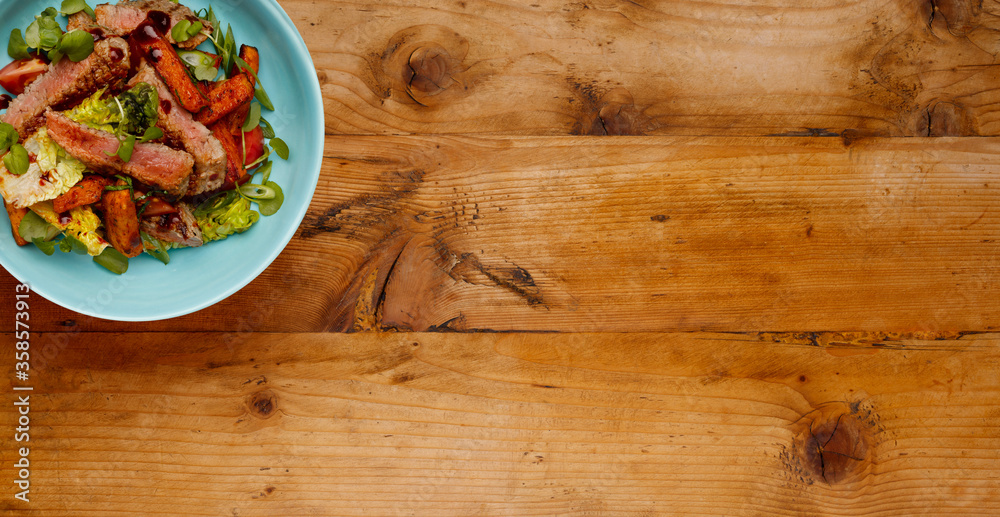 A delicious bowl of BBQ rump steak strips, sweet potatoe chips and vegetables, in a light blue bowl shot on a wooden background A delicious bowl of BBQ rump steak strips, sweet potatoe chips and vegetables, in a light blue bowl shot on a wooden background
