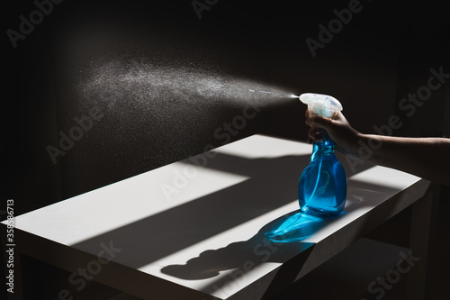 Crop anonymous person pushing dispenser mechanism and directing white cleaner flow on black wall while bottle being placed on white table with shadows