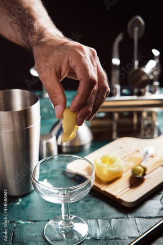 Crop anonymous male bartender decorating empty coupe glass with fresh lemon zest while preparing cocktail at bar counter