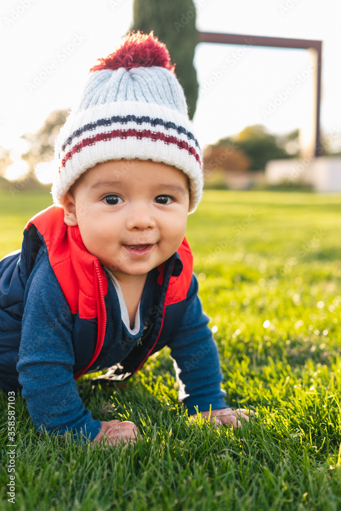 Cute ethnic toddler in warm clothes smiling and looking at camera while crawling on green meadow during family weekend in countryside Cute ethnic toddler in warm clothes smiling and looking at camera while crawling on green meadow during family weekend in countryside