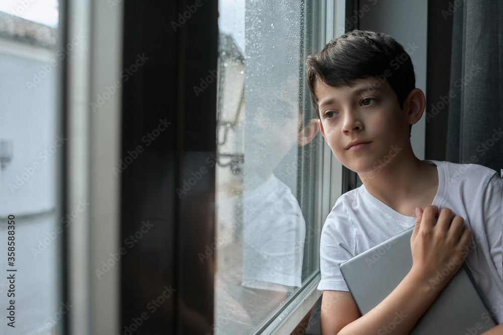 Thoughtful child in casual t shirt standing with tablet and leaning on window while observing rain in street Thoughtful child in casual t shirt standing with tablet and leaning on window while observing rain in street