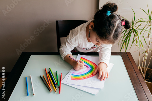 From above of anonymous girl in casual clothes sitting on chair at wooden table and drawing colorful rainbow with pencil on paper photo