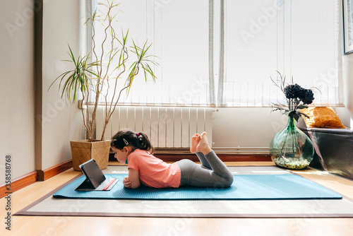 Side view of positive little girl lying down on mat near window at home resting after practicing yoga throughout video tutorials on tablet