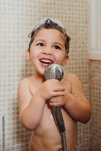 Smiling little child with foam on head standing in bathroom with shower and singing while looking away photo