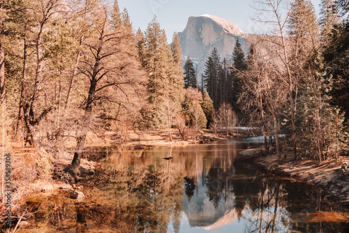 Spectacular peaceful landscape of calm river flowing through coniferous forest in mountain valley with rocky cliff reflected in sunny spring day in Yosemite National Park in California