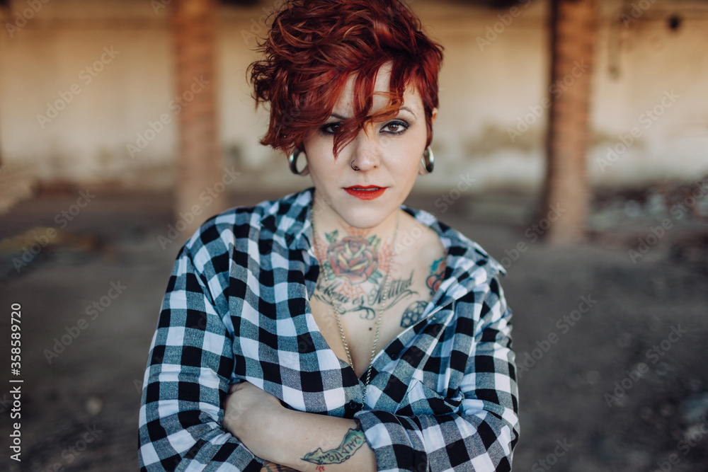 Independent stylish young female with trendy haircut and tattoos wearing casual checkered shirt looking at camera while standing against blurred shabby building