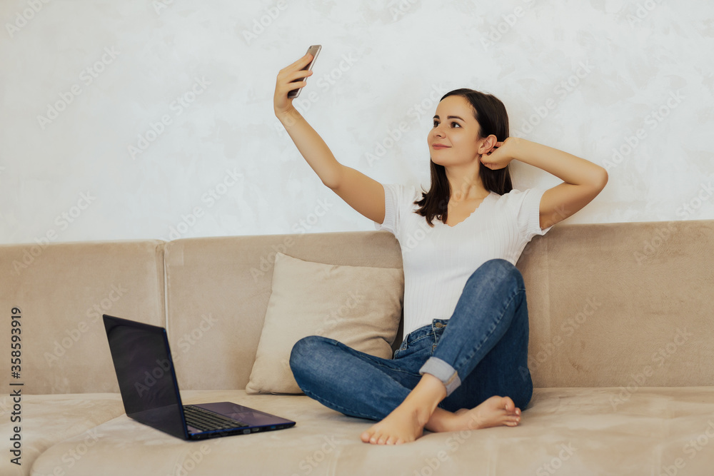 Smiling woman is sitting on a sofa in a living room with a phone. A young girl takes a selfie. Rest break. Happy woman sitting on the couch and using mobile phone for selfie or video chat. Copy space. Smiling woman is sitting on a sofa in a living room with a phone. A young girl takes a selfie. Rest break. Happy woman sitting on the couch and using mobile phone for selfie or video chat. Copy space.