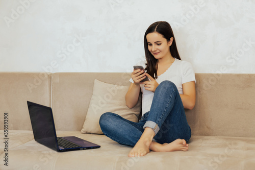 Young woman is sitting on the sofa in her own house with a phone happy talking to colleagues while working on the laptop, work from home. Copy space.