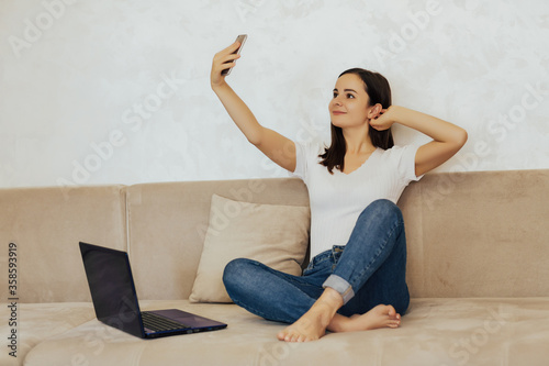 Smiling woman is sitting on a sofa in a living room with a phone. A young girl takes a selfie. Rest break. Happy woman sitting on the couch and using mobile phone for selfie or video chat. Copy space.