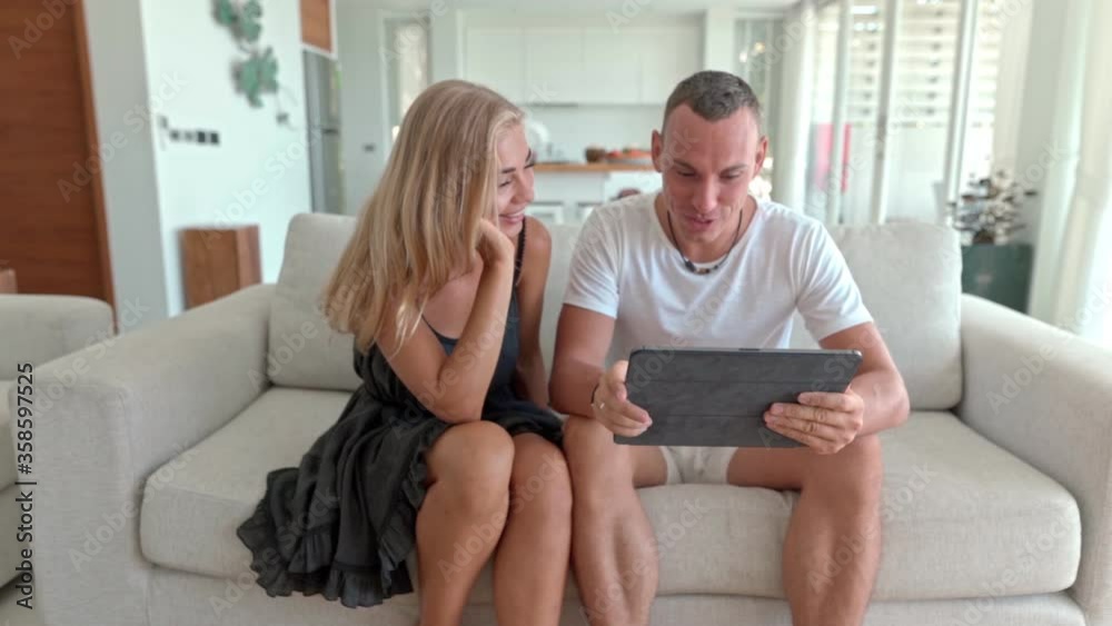 Happy young couple sitting on sofa, using tablet computer, browsing, laughing at funny photos, videos, having fun together, enjoying their weekend at home, 4K shot Happy young couple sitting on sofa, using tablet computer, browsing, laughing at funny photos, videos, having fun together, enjoying their weekend at home, 4K shot