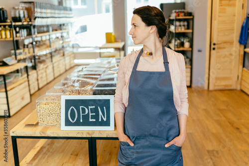 Mixed race woman worker or store owner with standing in the doorway of her grocery shop looking aside smiling. Portrait of girl assistant wearing apron welcoming inviting open door of store.