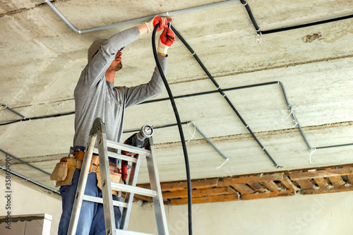 Worker electrician standing on aluminium ladder installing electrical cables laid in protective corrugation installed on the ceiling and wall in a room under construction. photo