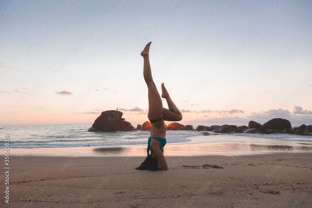 Young attractive latin woman wearing a bikini practicing yoga on the beach at sunset, healthy mental and body lifestyle concept.