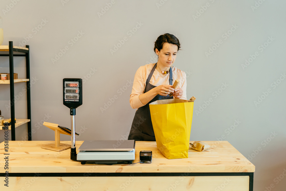 Female bakery worker behind the counter ready to service. Seller of zero waste shop on background of fresh organic homemade bread on wooden shelves in plastic free store. Owner small local business. Female bakery worker behind the counter ready to service. Seller of zero waste shop on background of fresh organic homemade bread on wooden shelves in plastic free store. Owner small local business.