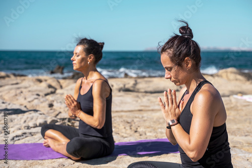 Two woman meditating on a rocky beach in sunny day in lotus pose