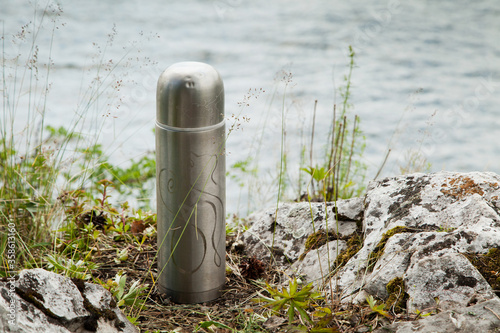 A tourist steel thermos with an engraving stands on the Bank of the river.