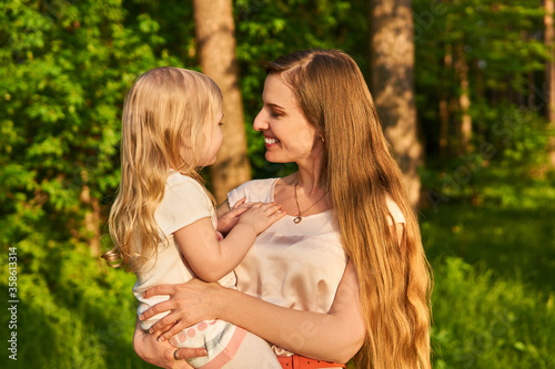 young woman affectionately looks at her little daughter, which she holds in her arms, against the background of nature