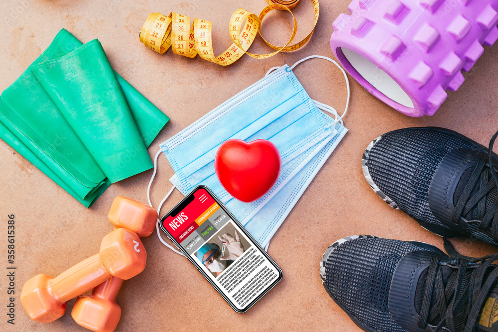 a top view of a black phone with headphones showing covid-19 news above a blue mask, measure tape, shoes and a roller for workout at home