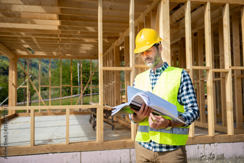 Engineer with hardhat and blueprints on building site of wood frame house under construction