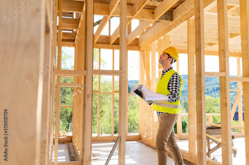 Obraz na plátně Engineer with hardhat and blueprints on building site of wood frame house under